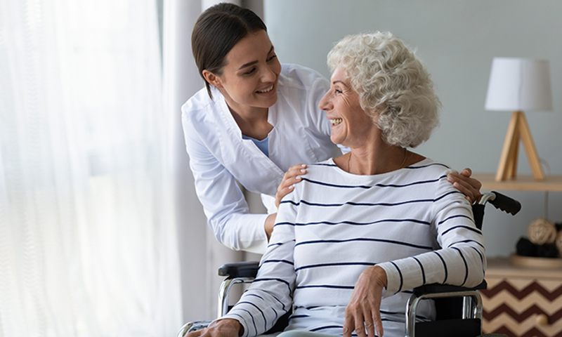 Caregiver smiling and supporting an elderly woman in a wheelchair at home.