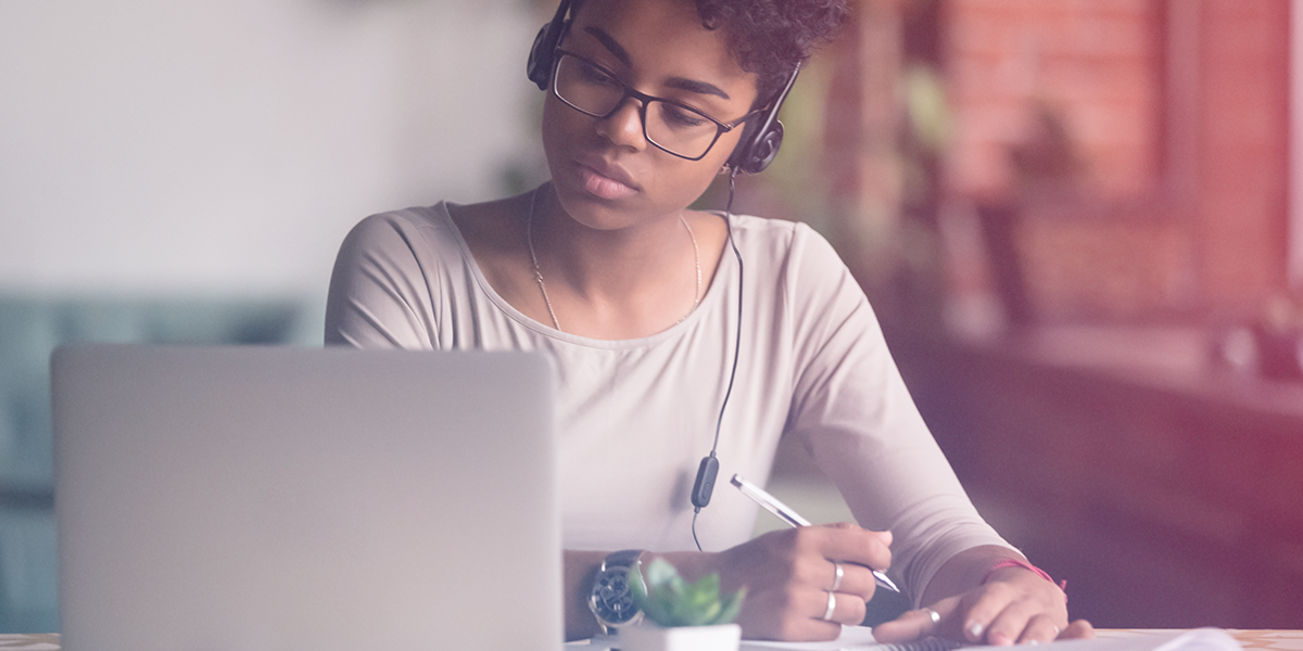 A young woman wearing headphones takes notes while looking at a laptop computer