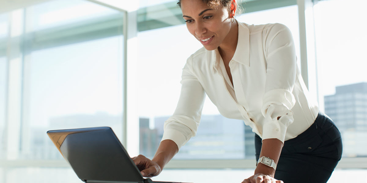 A woman in an urban office setting smiles as she leans over and works on a laptop computer.