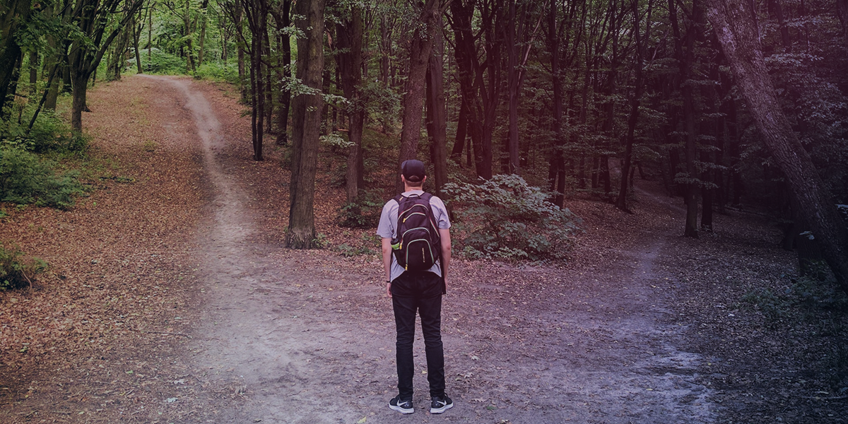 A man standing at a fork in the path in a forest. One path leads upward and is lighter, the other downward and darker.
