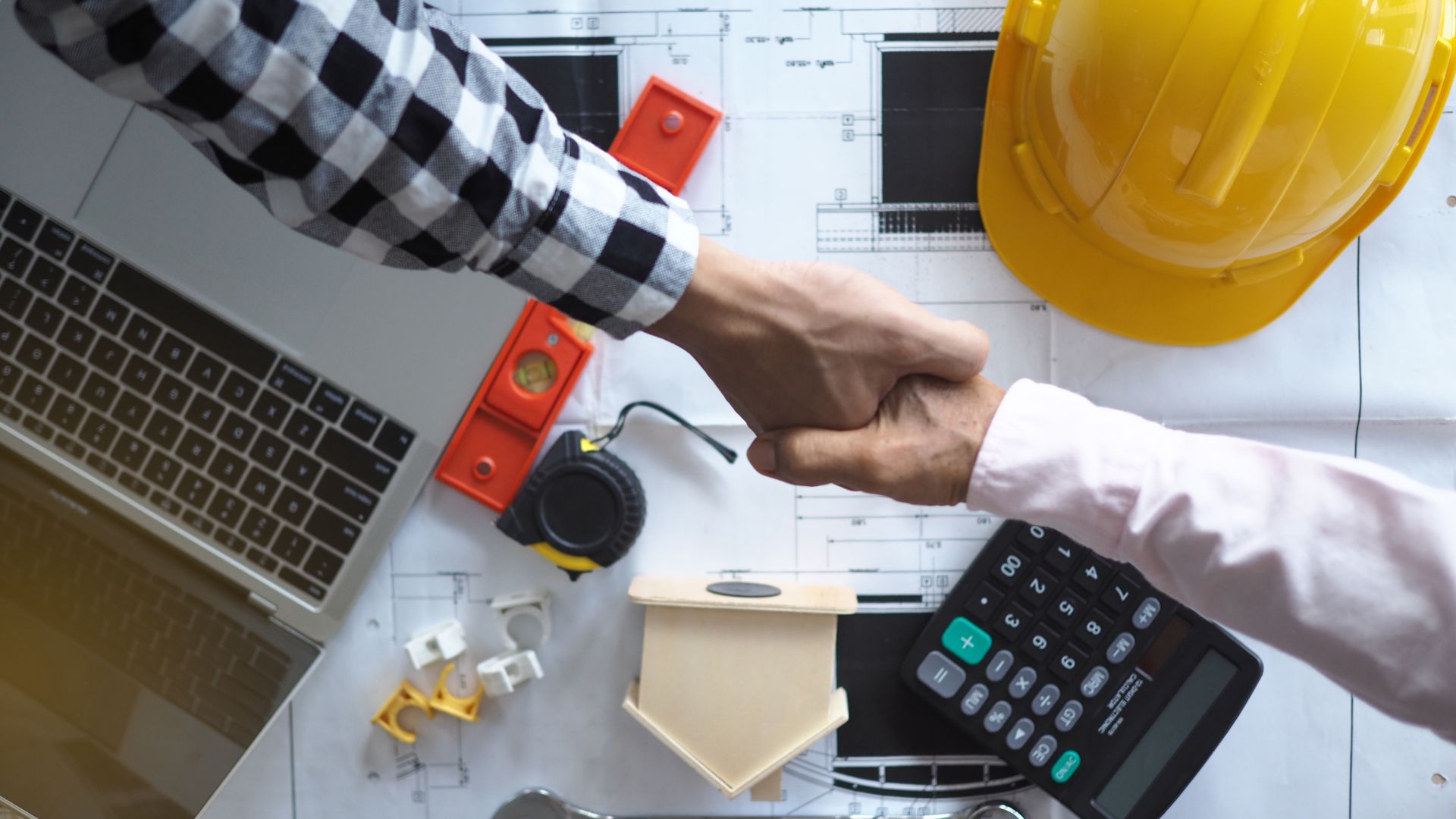 Two people shaking hands over a contract, hard hat, and calculator